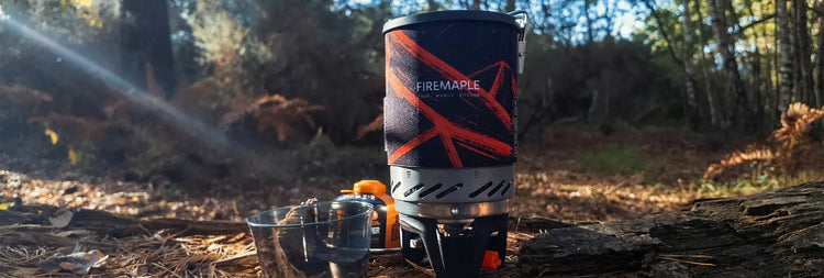 A black and orange Fire Maple camping mug with a matching fuel canister and two small cups, set up outdoors on a log in a sunlit forest.