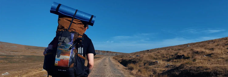 A hiker viewed from behind carrying a large brown and black backpack with a blue rolled sleeping pad on top, walking along a dirt path through dry grassy hills under a clear blue sky.